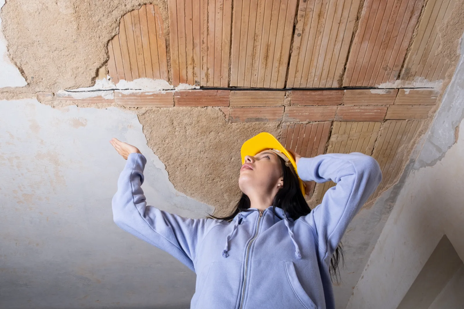 A young woman wearing a hard hat gazes up at a damaged ceiling.