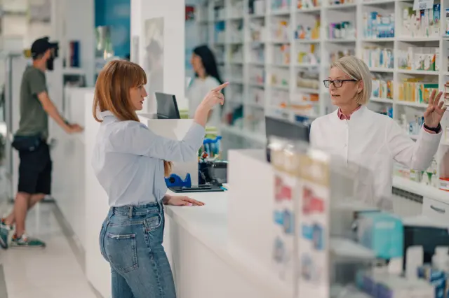 A pharmacist stands behind the counter while a customer asks for a specific medication.