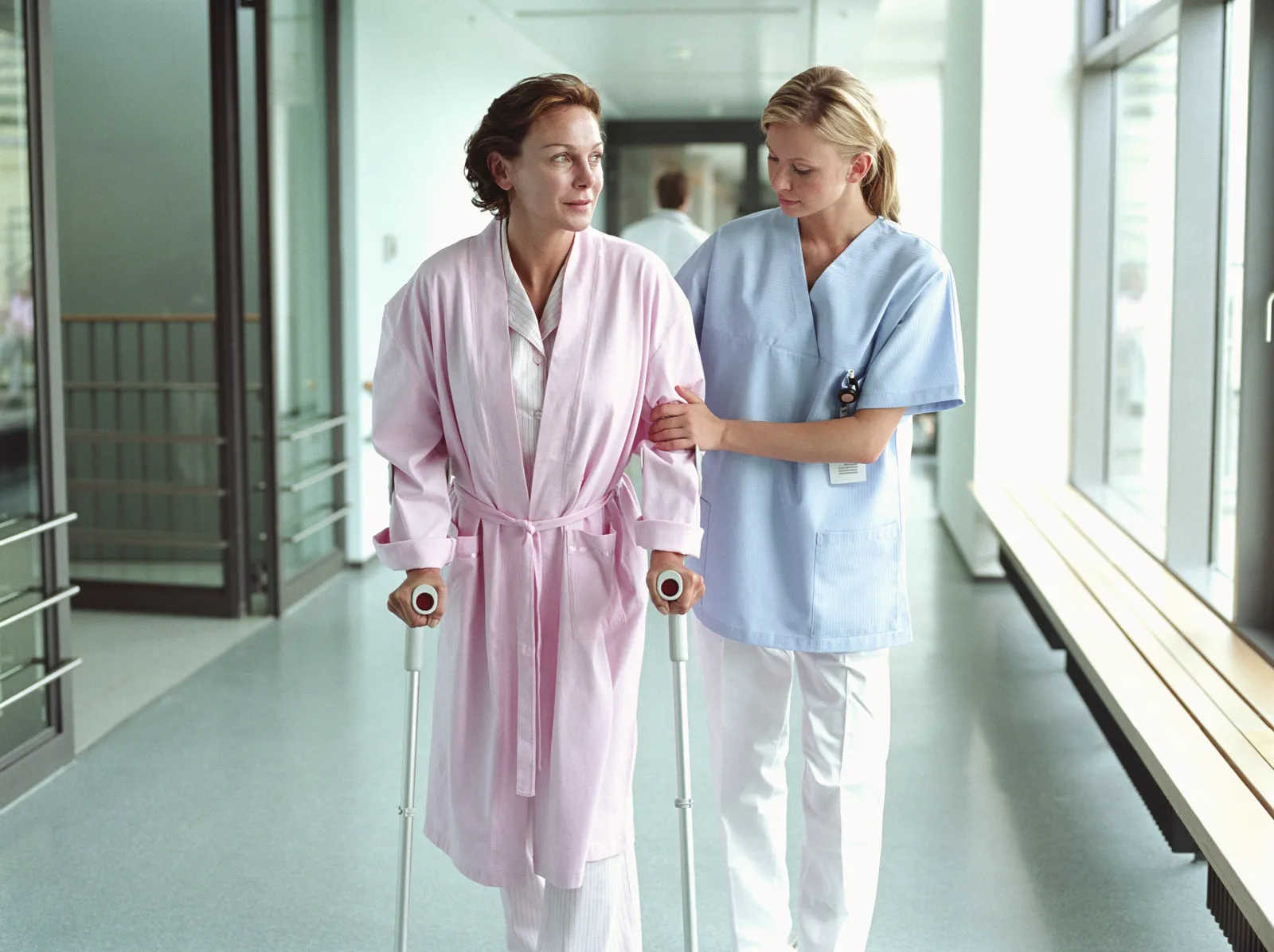 A nurse helps a patient on crutches walk in a hospital.