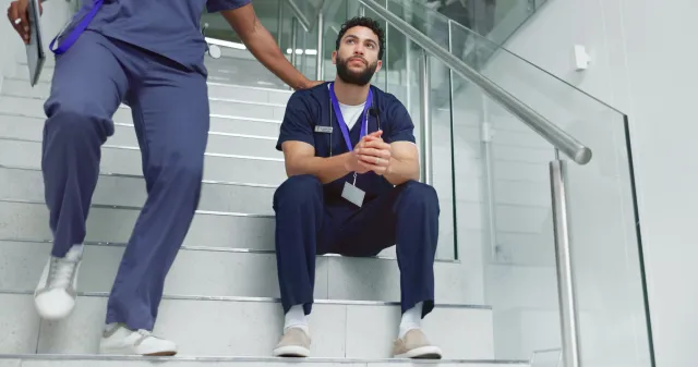 A male nurse sits on the stairs of a hospital appearing fatigued, while another walks past.