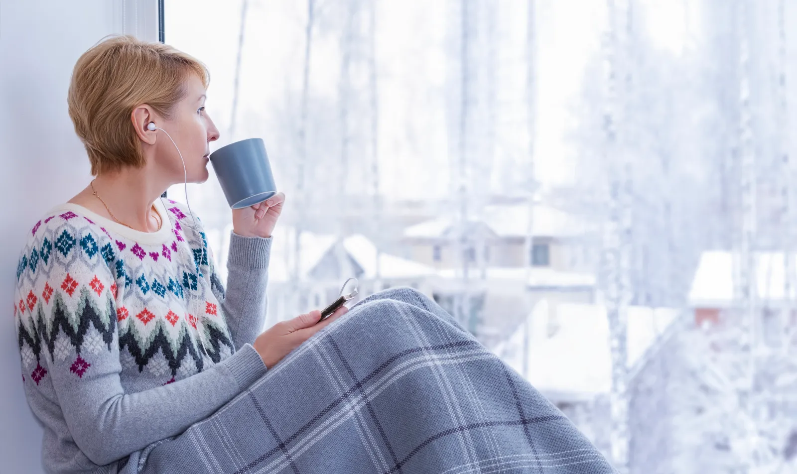 A woman covered in blanket by a window looking at snowy landscape outdoors
