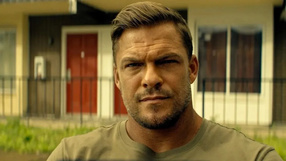 A man with a grumpy expression standing outside in a grey t-shirt.