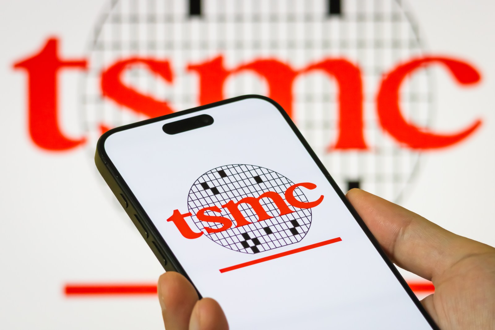 A person holds a smartphone displaying the logo of Taiwan Semiconductor Manufacturing Company, the world’s largest contract chipmaker, with the company’s logo visible on a screen in the background, on July 19, 2025, in Chongqing, China.