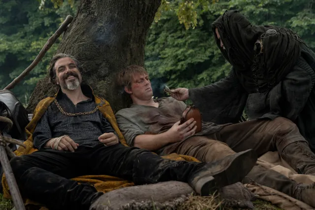 Two figures rest against a tree on a forest floor while a hooded figure leans in to offer one of them a drink.