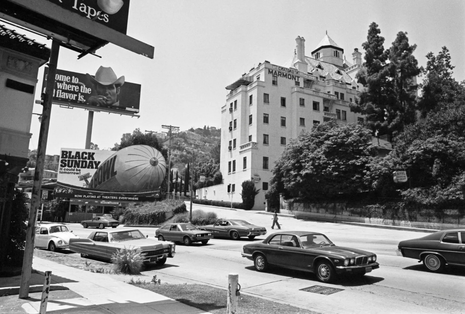 Traffic passes along Sunset Boulevard, passing beneath billboards advertising 'Black Sunday' and Marlboro cigarette with the Chateau Marmont hotel on the far side of the road, in Los Angeles, California, June 1977. Designed by architects Arnold A Weitzman and William Douglas Lee, the building is loosely based on the Chateau d'Amboise, a country house in the Loire Valley.