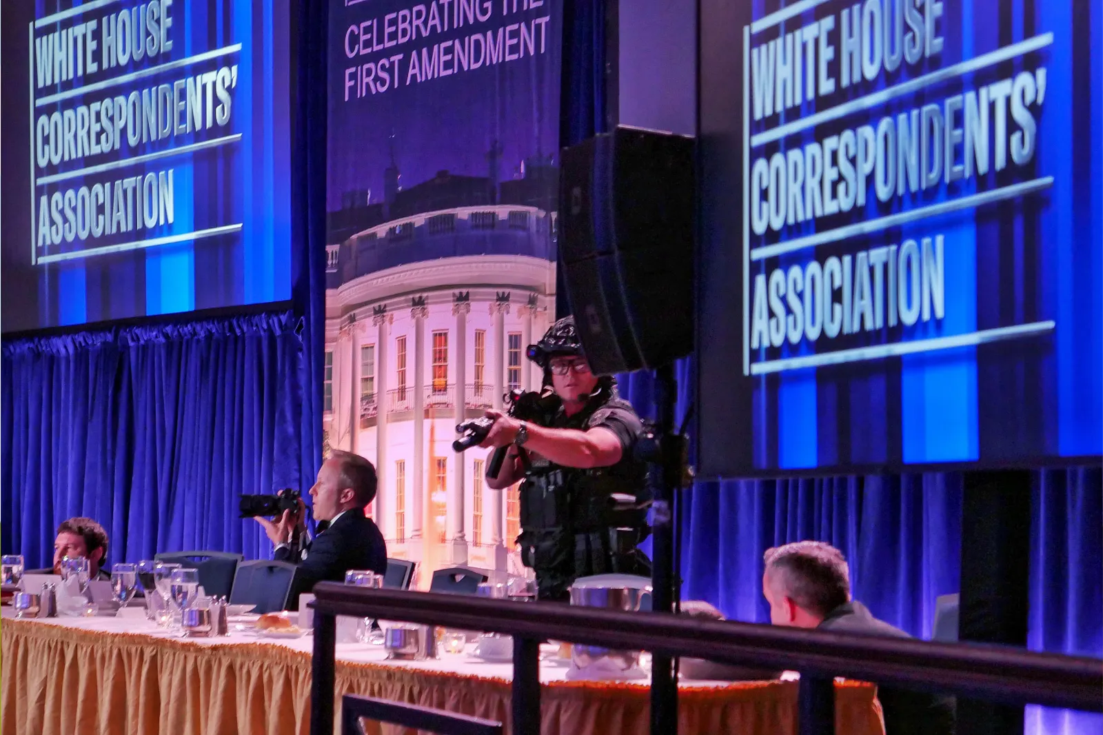 A photographer sits beside an agent with a gun, the White House Correspondents Association Dinner, April 25, 2026, Washington, D.C.