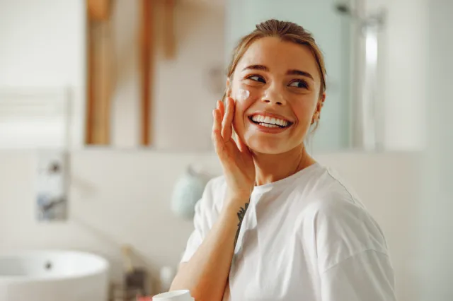 A stock image of a woman smiling and applying moisturizer.