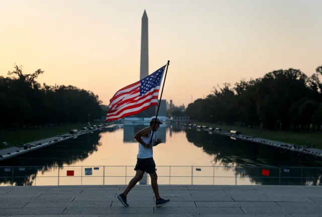 Pete Folch carries an American flag during a morning run past the Reflecting Pool as the city prepares for July 4th festivities on July 03, 2026, in Washington, D.C.