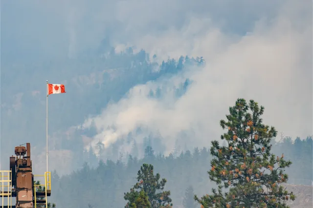 A Canadian flag blows in the high wind as the McDougall Creek wildfire burns above the City of West Kelowna, B.C. in August 18