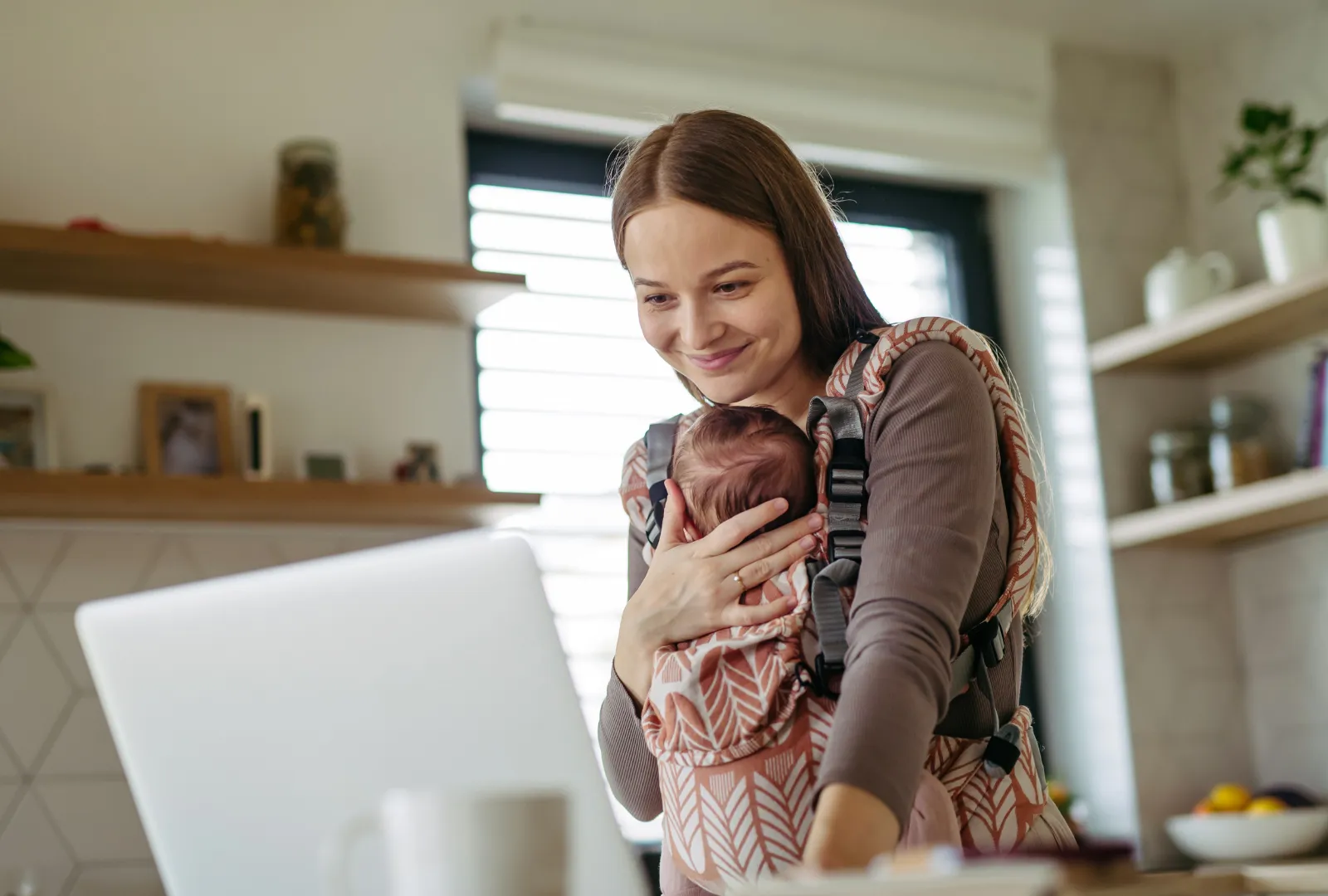 Mother taking online class while holding baby in ergonomic carrier.