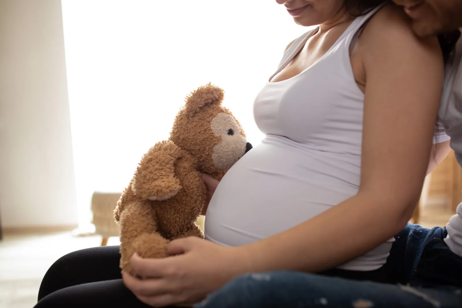 Stock Image: A pregnant woman and her partner sitting on a bed with a teddy bear.