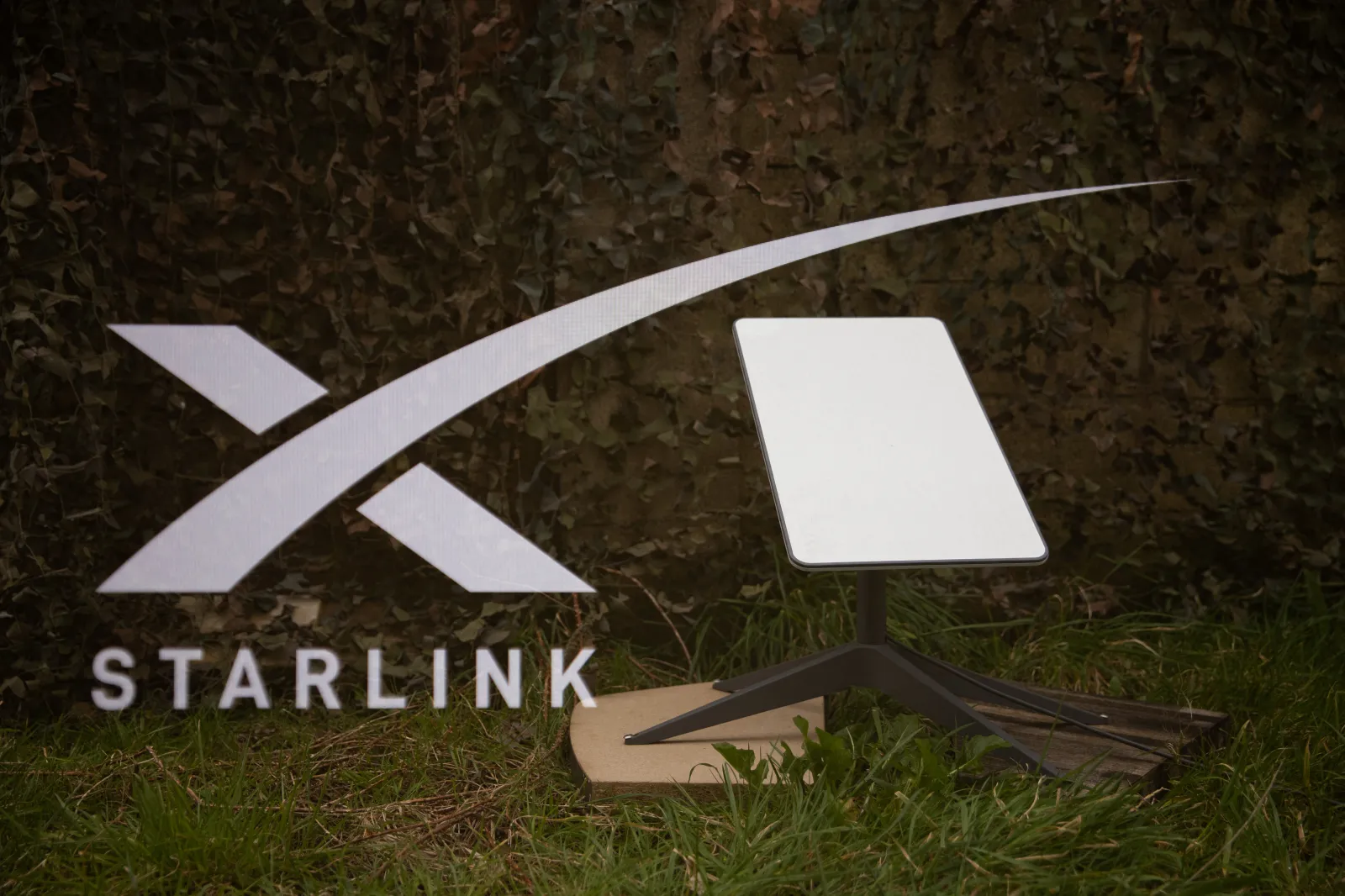 Illustration of the Starlink logo and satellite antenna on the ground in front of a military camouflage net in France on March 1, 2025.
