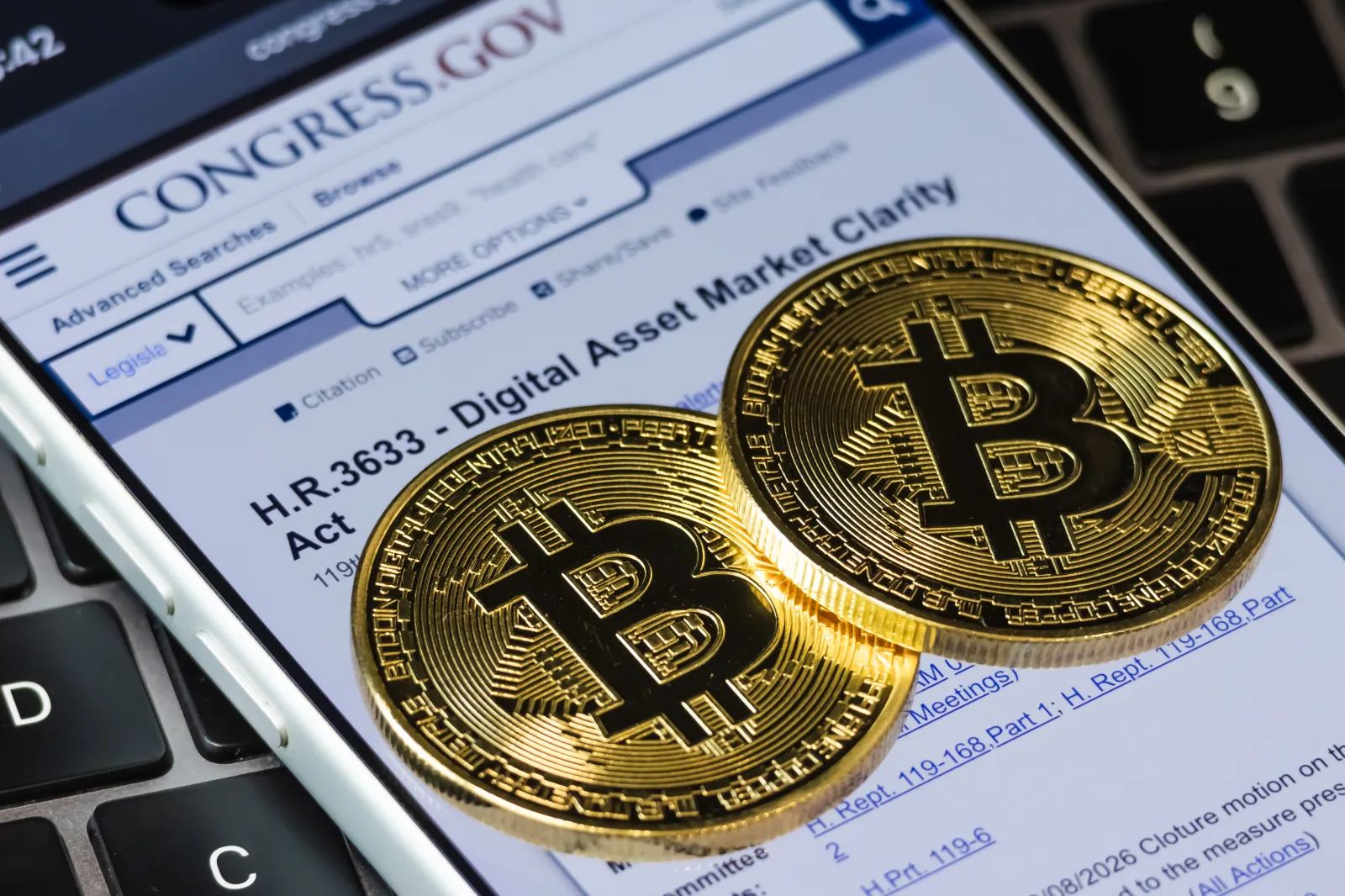 In this photo illustration, two gold-colored souvenir tokens bearing the Bitcoin symbol rest on an iPhone displaying the Congress.gov page for H.R. 3633, the Di