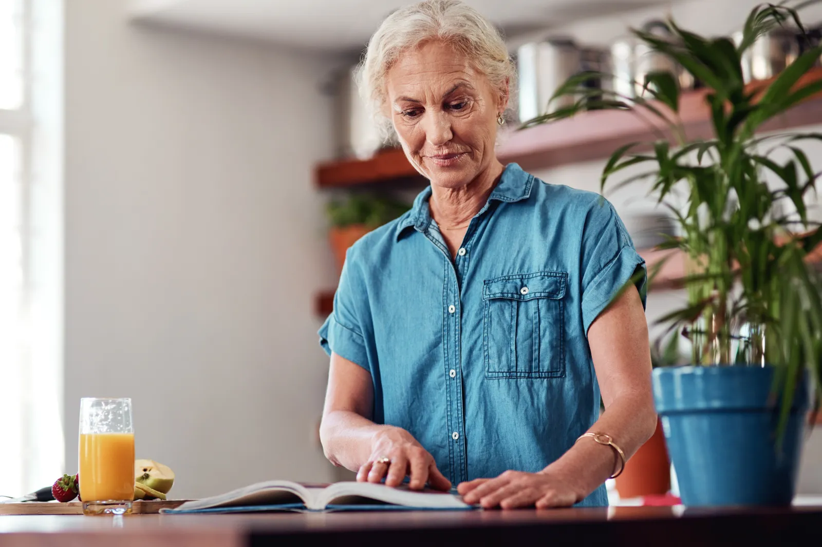 A stock photo of a senior woman reading from a recipe book while preparing breakfast in the kitchen.