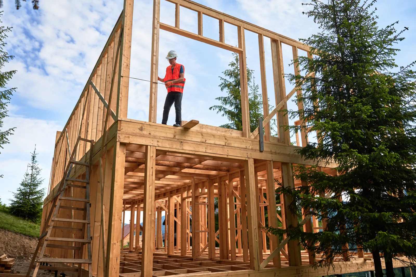 A worker using a tape measure while constructing a timber-framed two-story building.