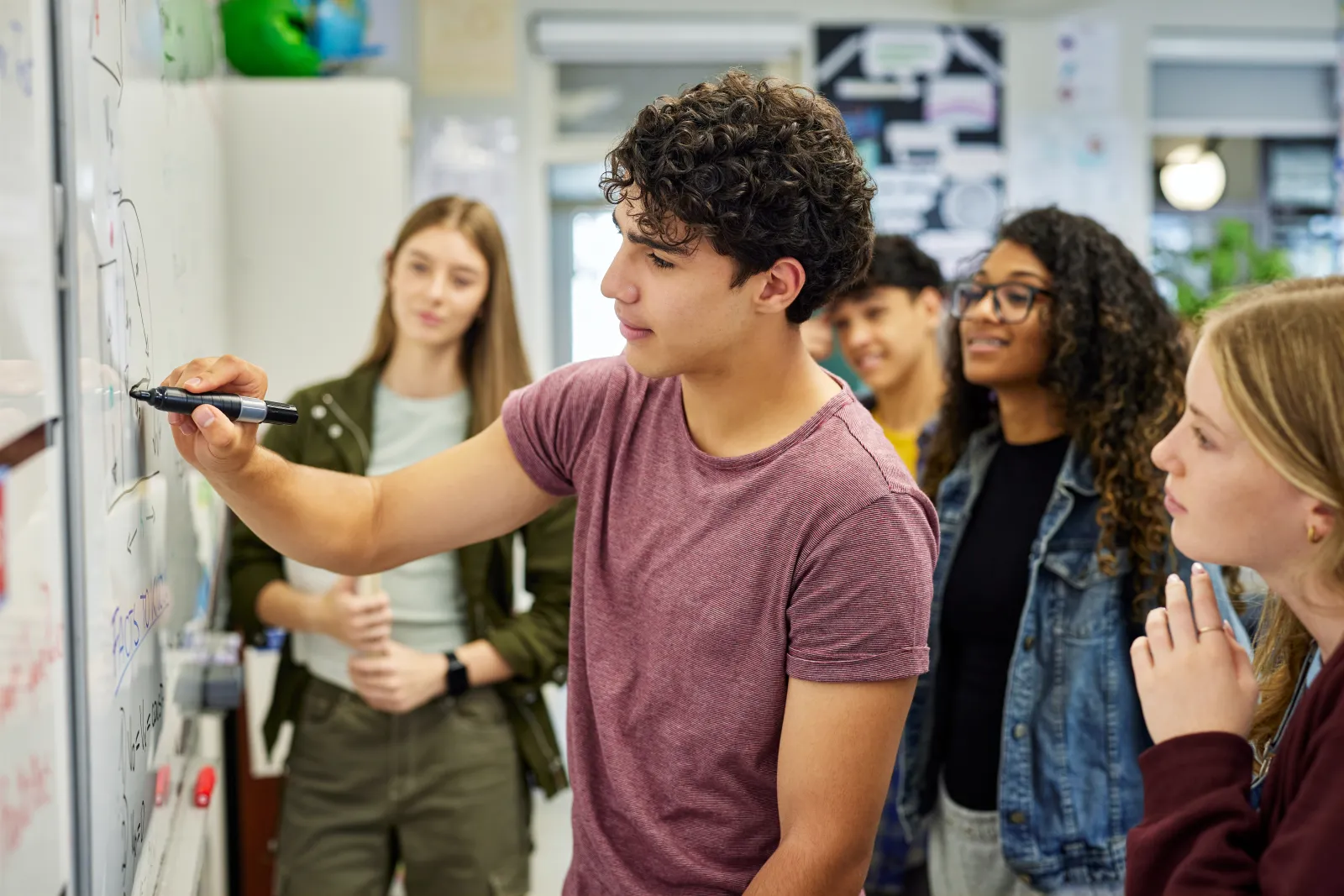A male student writes on whiteboard while explaining a concept to his classmates at school.