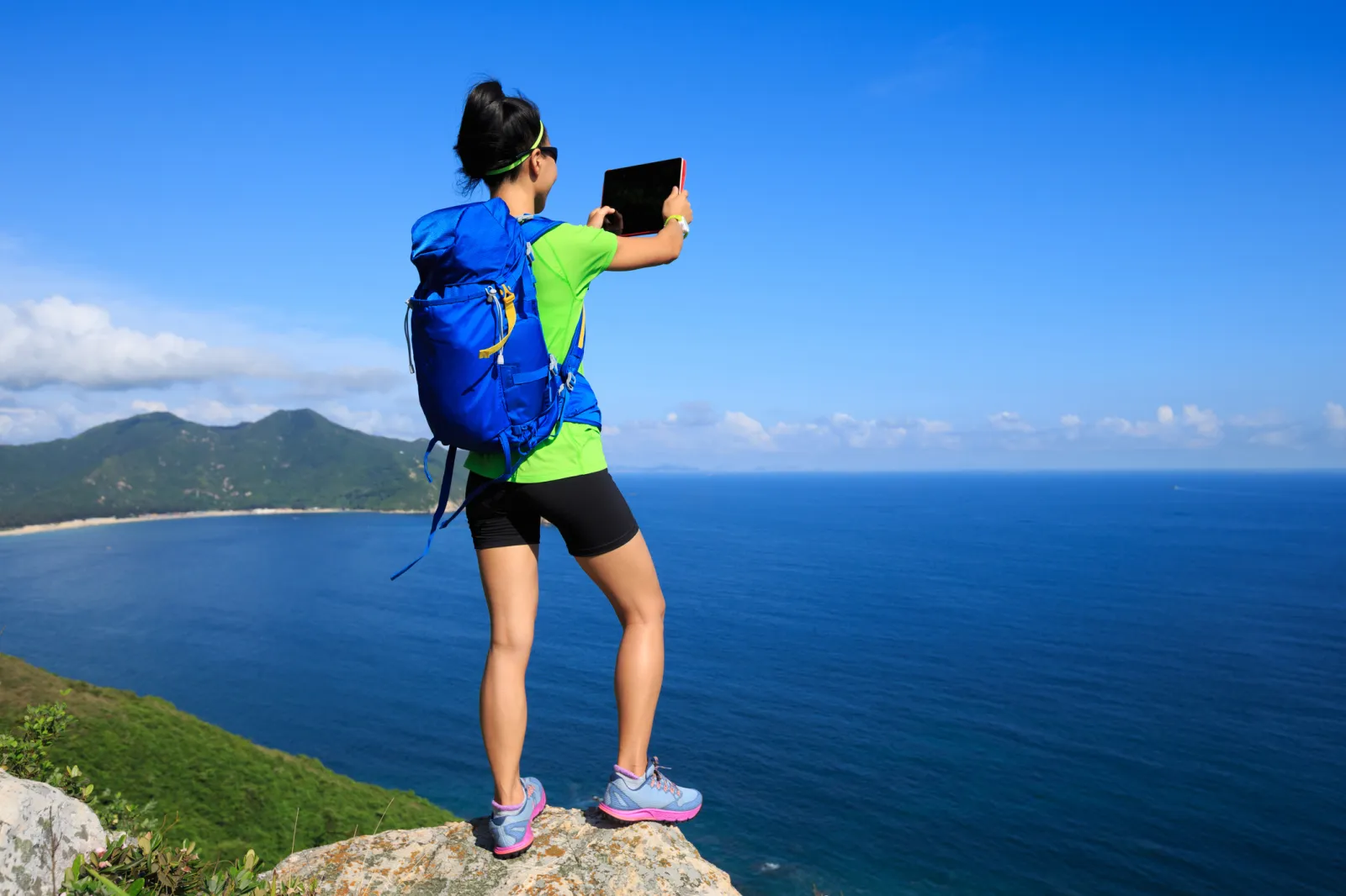 A hiker taking a picture of the sea with a tablet while standing at the edge of a rock cliff.